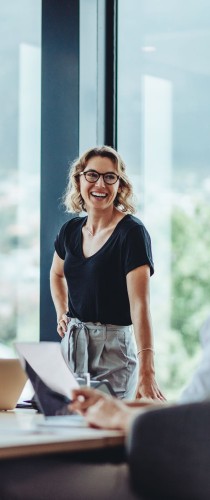 Woman with glasses smiling confidently in black top, standing by office window with urban backdrop.