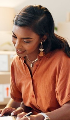 Woman in orange blouse with jewellery writing notes, focused on task. 