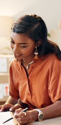 Woman in orange blouse with jewellery writing notes, focused on task. 
