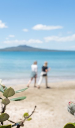 Two people standing on sandy beach with turquoise water and island in distance, framed by coastal plants