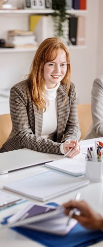 Woman with auburn hair in brown blazer smiling during professional meeting with organized workspace.