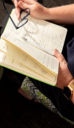 Hands holding glasses above journal with handwritten notes, gold watch visible on wrist.