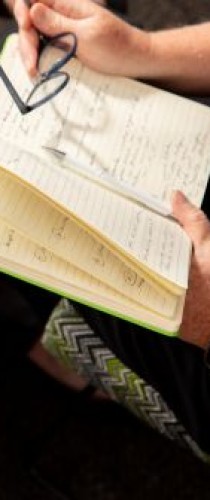Hands holding glasses above journal with handwritten notes, gold watch visible on wrist.