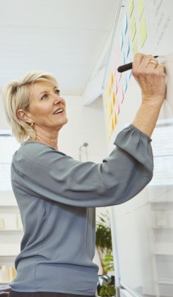 Woman in grey top writing on colorful wall chart, looking up thoughtfully during planning session.