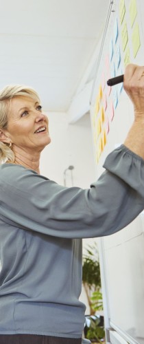 Woman in grey top writing on colorful wall chart, looking up thoughtfully during planning session.
