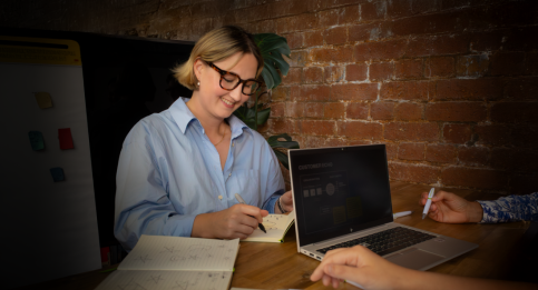 Team member taking notes during customer feedback session with laptop displaying dashboard against brick background. 