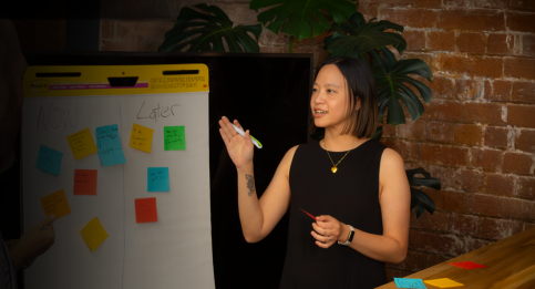 Workshop facilitator organising colour-coded sticky notes on whiteboard into 'Now vs Later' prioritisation framework. 