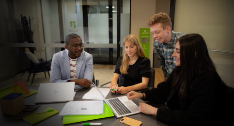 Diverse team of four professionals reviewing documents around a table, collaborating on project materials with laptops and branded stationery. 