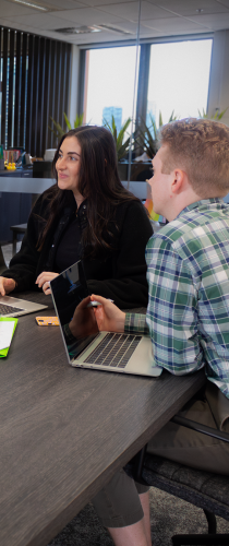 Two colleagues engaged in discussion at a meeting table with laptops, woman in black smiling while man in checkered shirt takes notes. 