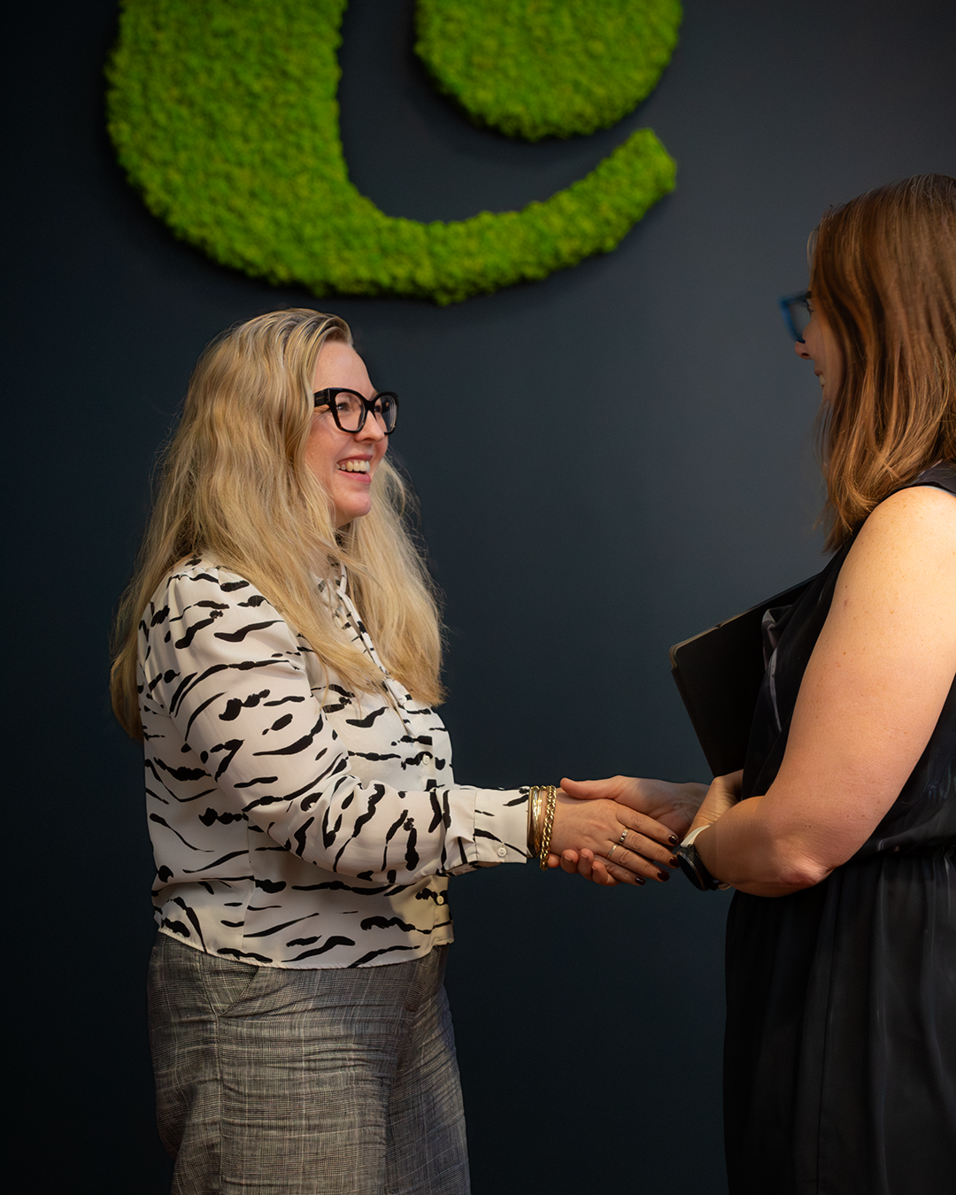 Colleagues sharing a warm handshake, one wearing patterned blouse and glasses. Contemporary office space features dark blue wall with distinctive green moss logo installation. 