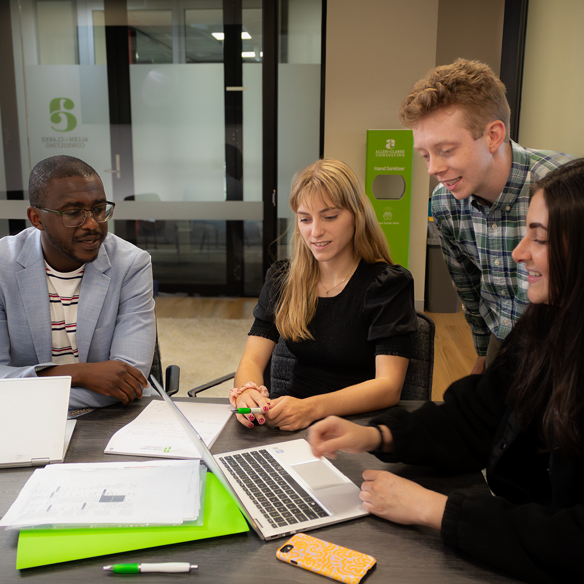 Diverse team of four professionals reviewing documents around a table, collaborating on project materials with laptops and branded stationery. 