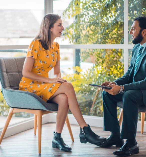 Professional consultation in bright setting with natural greenery backdrop, woman in yellow dress discussing with man in suit holding tablet.