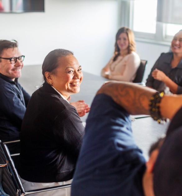 Diverse professionals engaged in a positive meeting environment, with a smiling woman in black jacket turning toward camera.