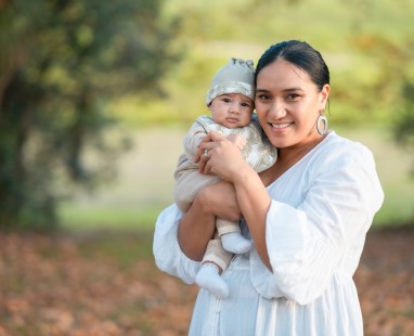 Māori woman in white dress smiling while holding pepi in patterned outfit, photographed outdoors. 