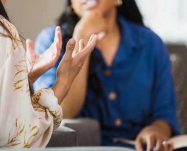 Close-up of a conversation between two people - one person has hands gesturing wearing patterned white top, while the other person wears a blue shirt. (Thumbnail)