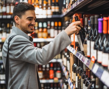 Smiling man in grey blazer selecting bottle from well-stocked shelves, examining label thoughtfully in retail environment.