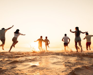 Silhouettes of diverse group joyfully running and splashing into water at sunset, creating dynamic movement and community spirit.