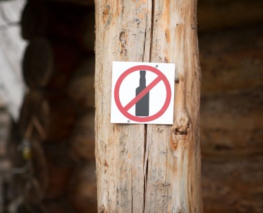 "No alcohol" prohibition sign mounted on weathered wooden post, featuring clear red circle-slash symbol over bottle silhouette against white background.