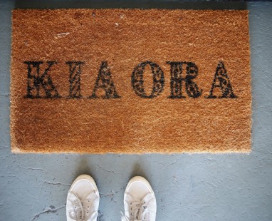 Brown doormat with Kia Ora greeting text on grey doorstop with white shoes 