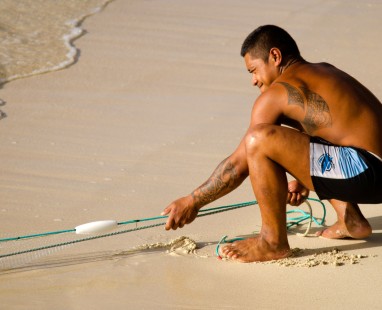Person crouched on beach drawing lines in sand with stick near ocean waves