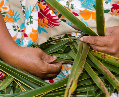 Hands weaving traditional flax basket with colorful tropical patterned fabric in background