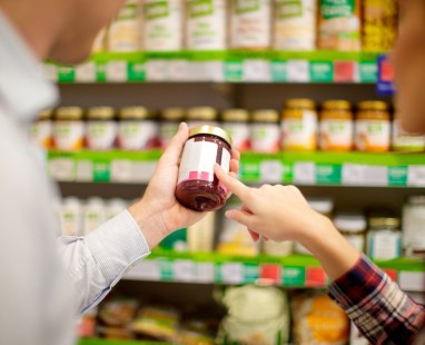Person examining product label on jar while shopping in grocery store aisle with shelves of packaged goods