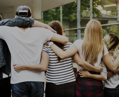 Group of young people with arms around each other's shoulders, viewed from behind, demonstrating unity and collaboration