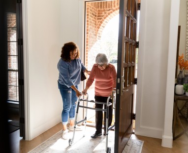 Carer with elderly person using walker in hallwa