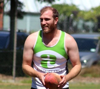 Team member in green and white Allen + Clarke singlet holding a rugby ball on a grass field. 
