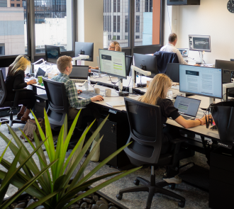 Collaborative team works at dynamic open-plan office with city views, multiple monitors displaying documents, and indoor plants adding vibrant greenery.