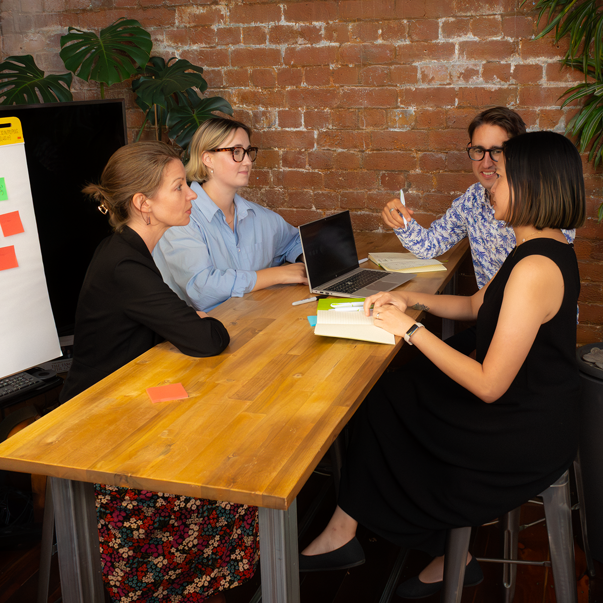 Four colleagues engaged in animated discussion around a table with laptop and notebooks. Exposed brick wall and plants create a warm, creative workspace with whiteboard visible nearby. 