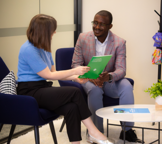 Two professions sharing an Allen + Clarke document. Person in pink checkered blazer smiles while receiving the report. Blue chairs and small table create a comfortable consultation space.