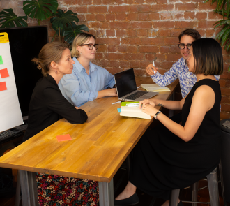 Four colleagues engaged in animated discussion around a table with laptop and notebooks. Exposed brick wall and plants create a warm, creative workspace with whiteboard visible nearby. 