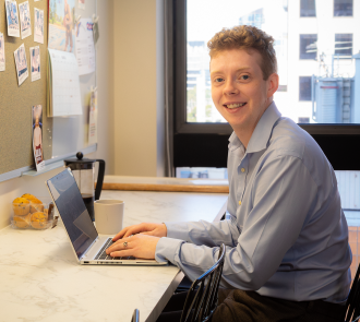 Smiling professional in light blue shirt working on laptop. Bulletin board with photos nearby, coffee cup and muffins on counter, city view through window in bright office space. 