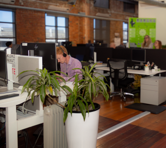Open plan workspace with exposed brick walls. Team member in purple checked shirt wearing headphones focuses on computer. 