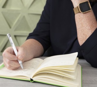 Person taking notes in a bound journal with white pen, gold smartwatch visible against navy blue sleeve