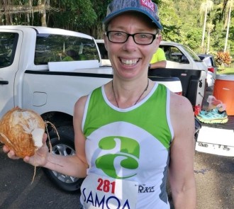 Team member in Allen + Clarke singlet holding a coconut at Samoa Events race, smiling in front of tropical foliage..