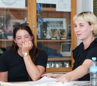 Two female colleagues reviewing documents together at a table, with awards displayed in a wooden cabinet behind them. 