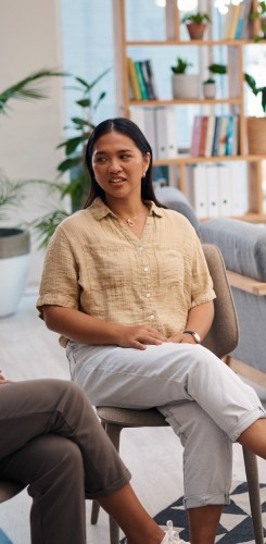 Woman smiling while seated in modern office space with wooden shelving unit displaying books and plants, wearing beige top and white pants.