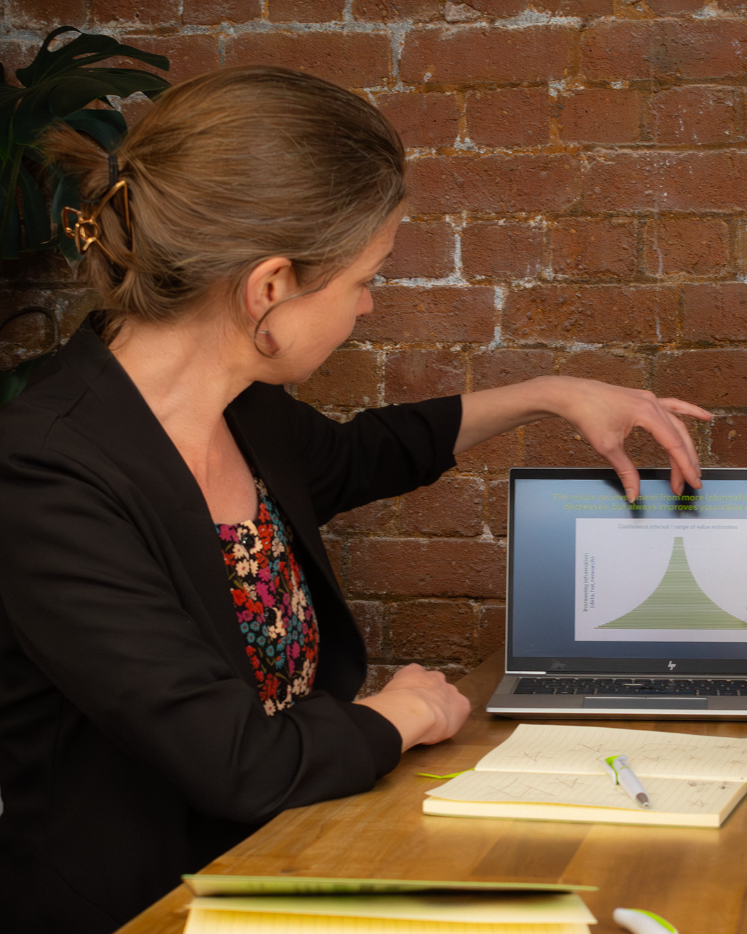 Professional in black blazer analyzing data on laptop, working with reference materials at wooden desk against brick wall backdrop.