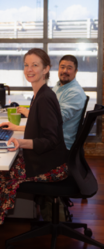 Two colleagues working together at adjacent desks with computers in bright office space with large windows.