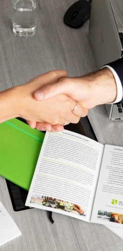 Business handshake over open documents and laptop, signifying partnership agreement with Allen + Clarke branded materials visible.