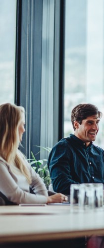 Two professionals engaged in discussion at meeting table near floor-to-ceiling windows, creating collaborative atmosphere in bright space.