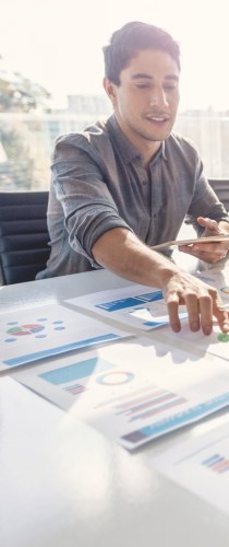 Team member presenting data analysis with charts and reports spread across table, gesturing to emphasize key insights.