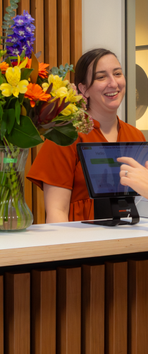 Staff member in orange shirt smiling at reception desk with digital tablet, beside vibrant floral arrangement of purple and yellow blooms.