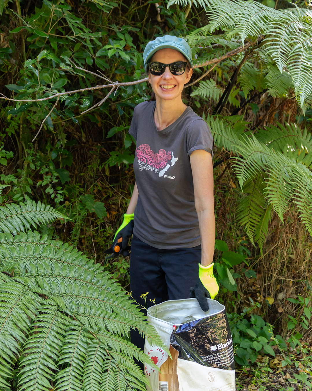 Smiling woman in grey cap and sunglasses kneeling among lush native ferns doing conservation work.  