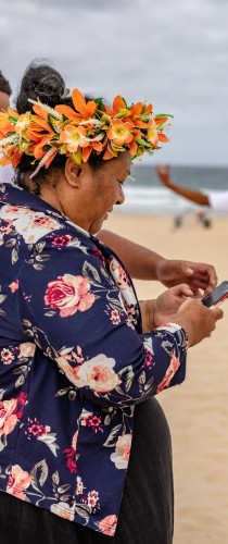 Participant wearing flower crown and floral shirt checking mobile device at beachside event. 