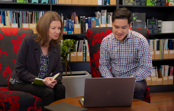 Allen + Clarke consultants collaborate in cozy library space, sharing insights over laptop while surrounded by policy resources and branded publications.