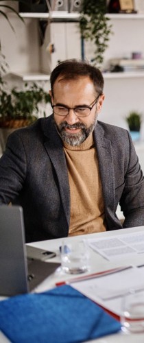 Team member reviewing documents at desk with laptop, wearing smart casual attire in plant-adorned workspace.