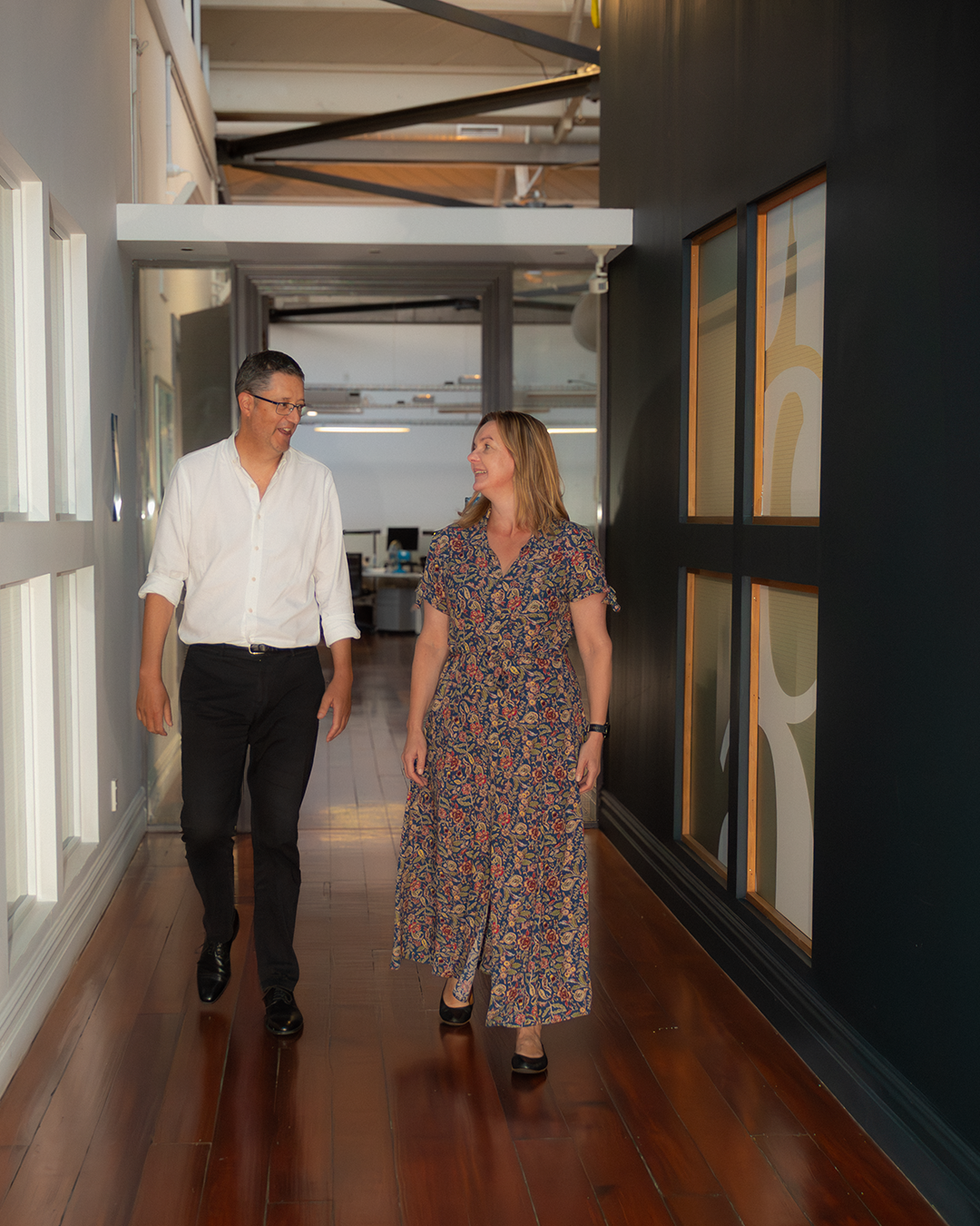 Two colleagues walking through office corridor with framed displays, engaged in professional conversation while navigating workspace.